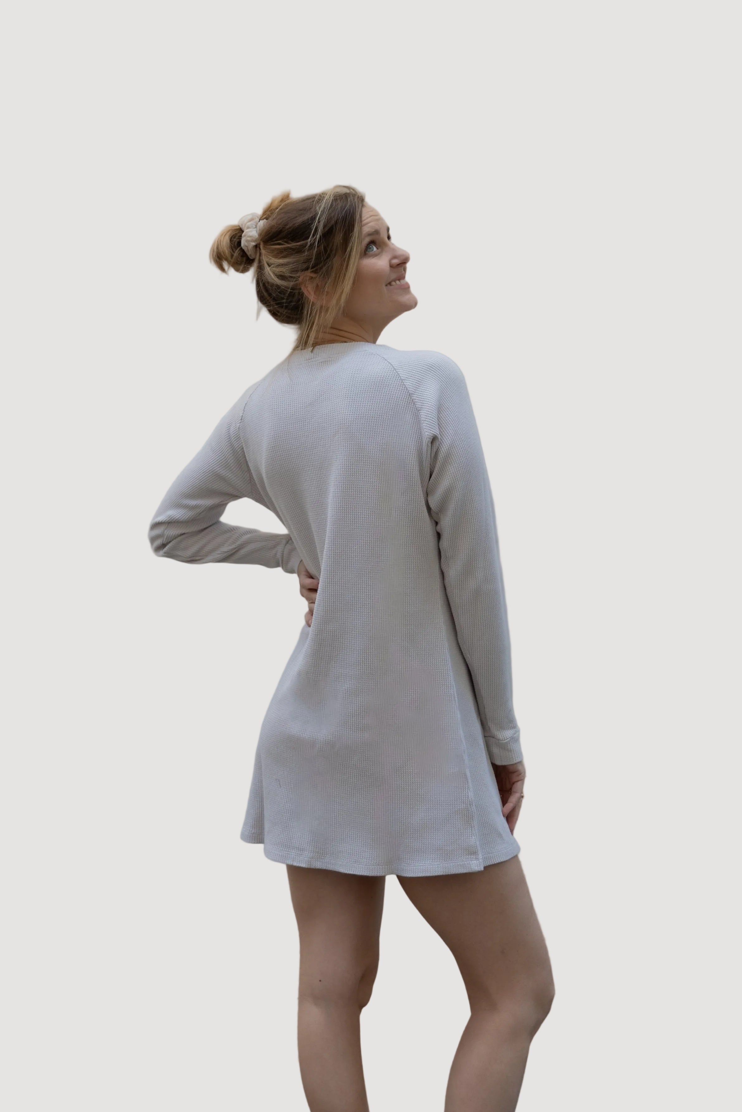 A woman wearing a white, long-sleeved, knee-length dress, standing against a neutral background.