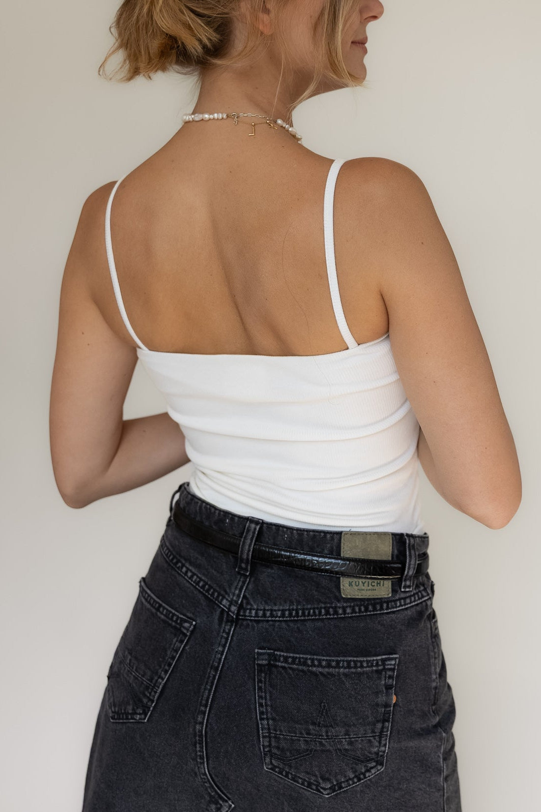 Woman wearing a white tank top and dark jeans against a plain background