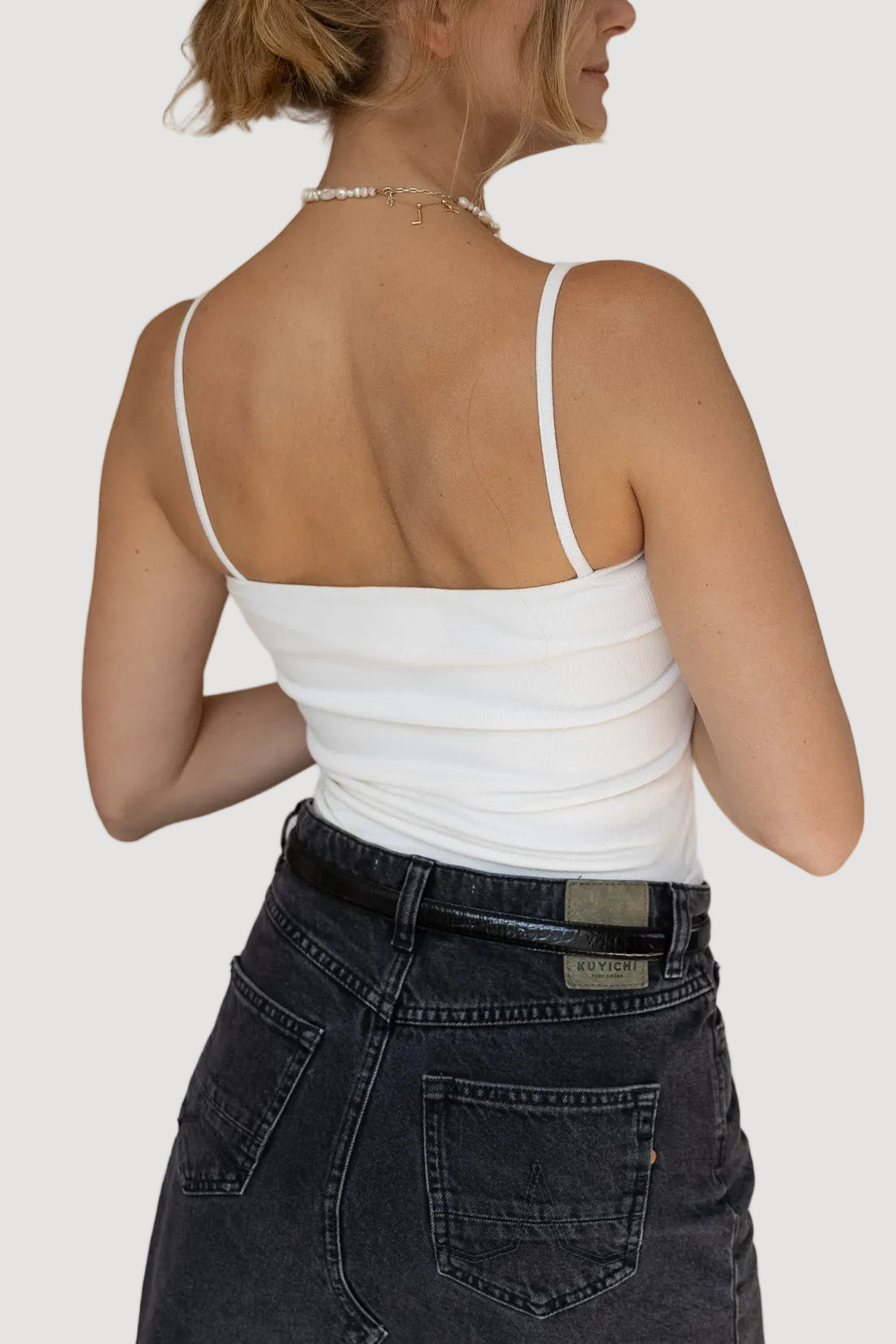 Woman wearing a white tank top and dark jeans against a plain background