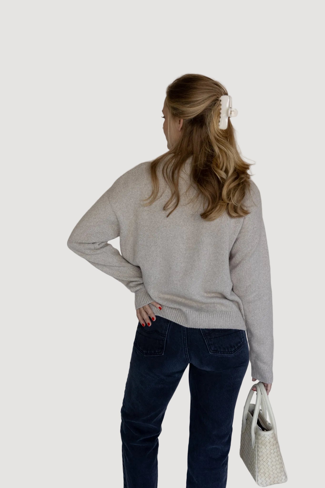 Woman wearing a gray sweater and dark blue jeans, standing against a plain white wall.