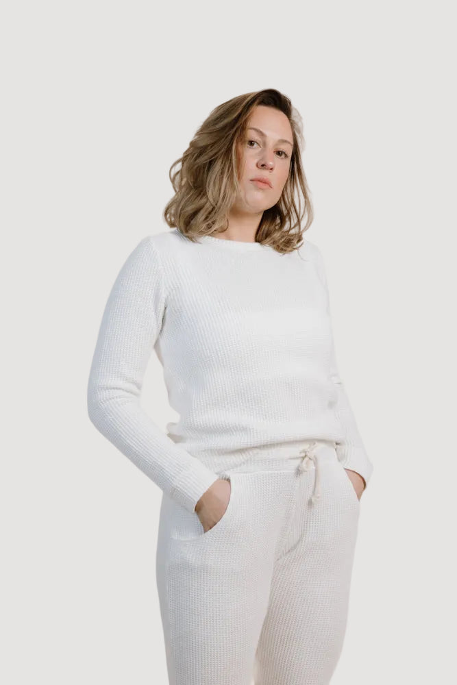 A person wearing a white long-sleeved kali sweater with round neckline, paired with white pants, standing with hands in pockets.