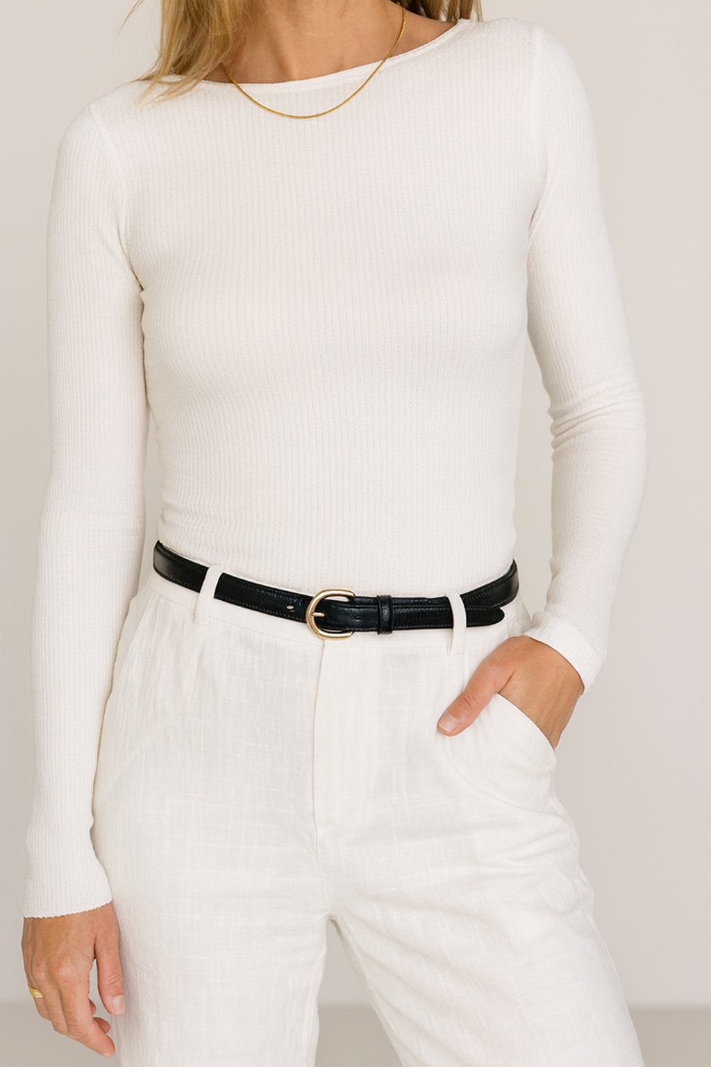 Person wearing a white long-sleeve top and white pants with a black belt against a plain background