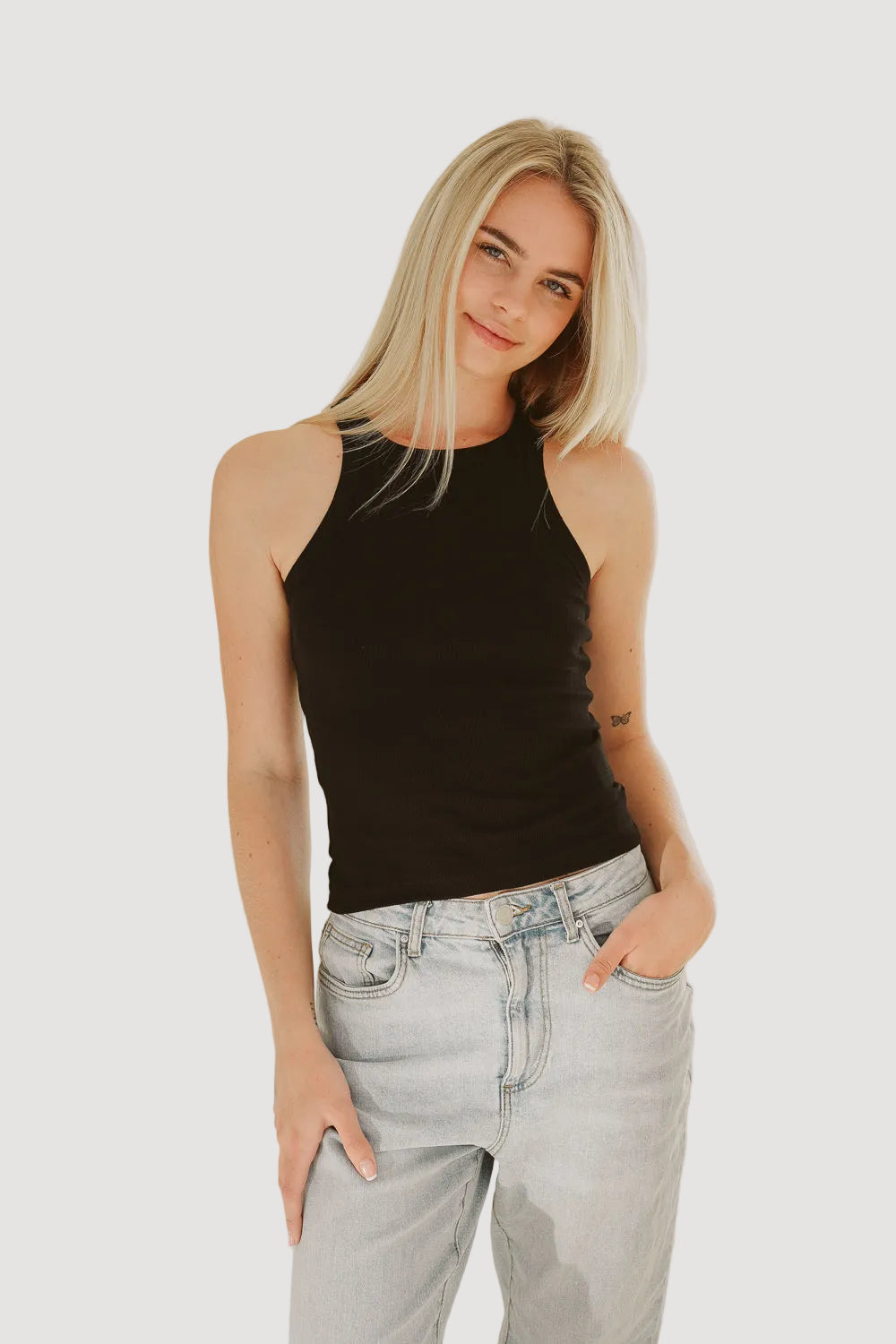 A woman wearing a black tank top and light blue denim jeans standing against a neutral background.