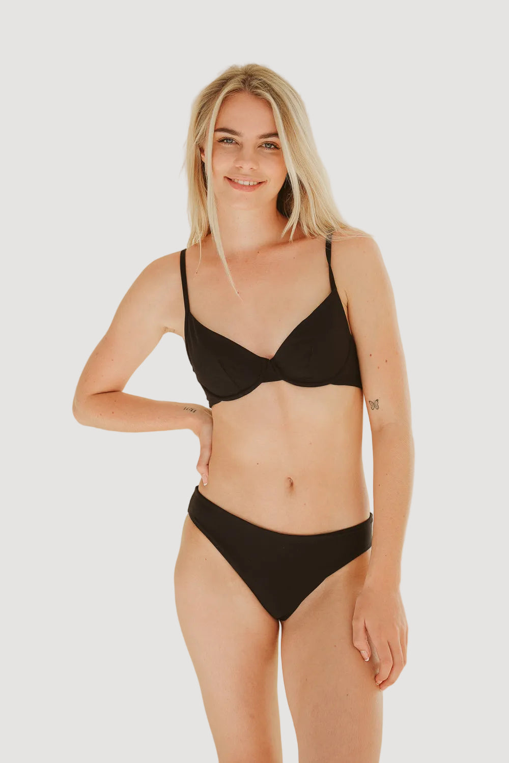A female model wearing a black medium coverage bikini top with underwire design, made from biobased fabric, posed against a beige background.