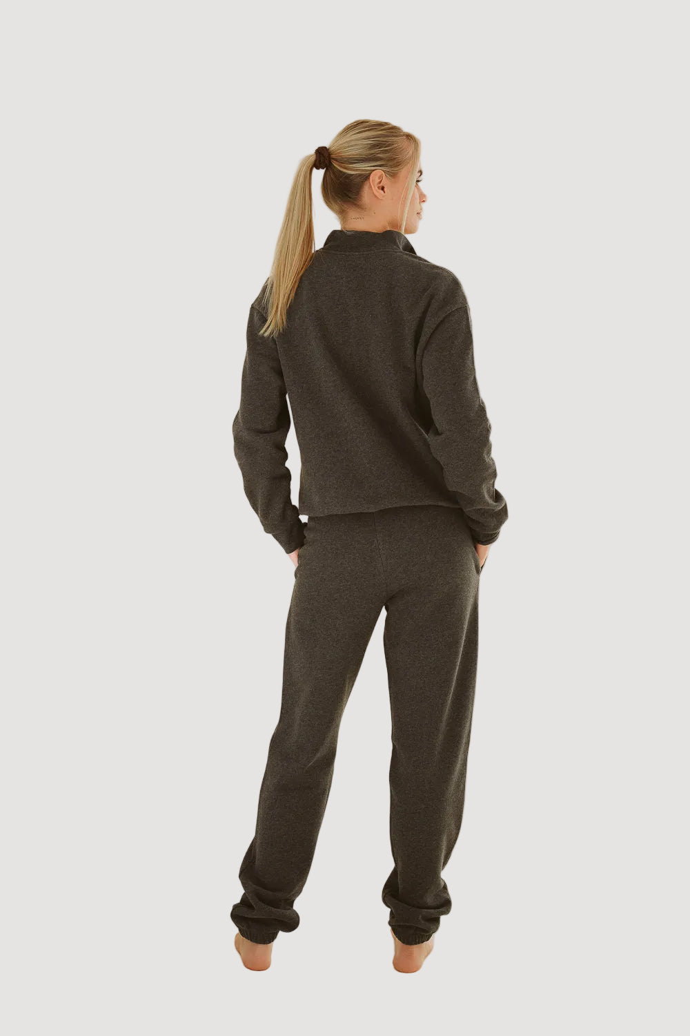 A person standing with their back to the camera, wearing dark grey baggy pants and a matching dark grey top, in a studio setting with a plain background.