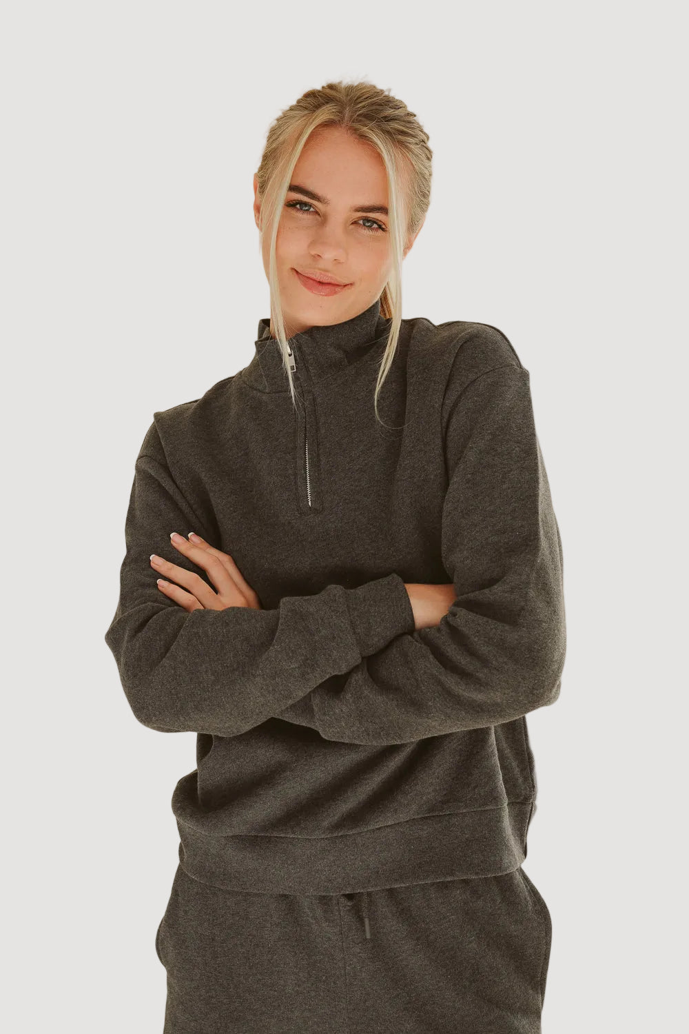 A woman wearing a dark grey half zip sweater over a white top, standing with her arms crossed.