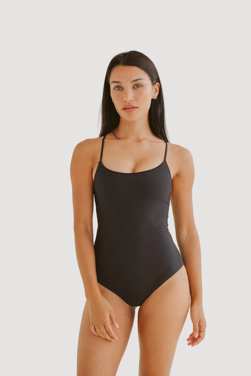 A woman wearing a black one-piece swimsuit with spaghetti straps, standing against a neutral background.