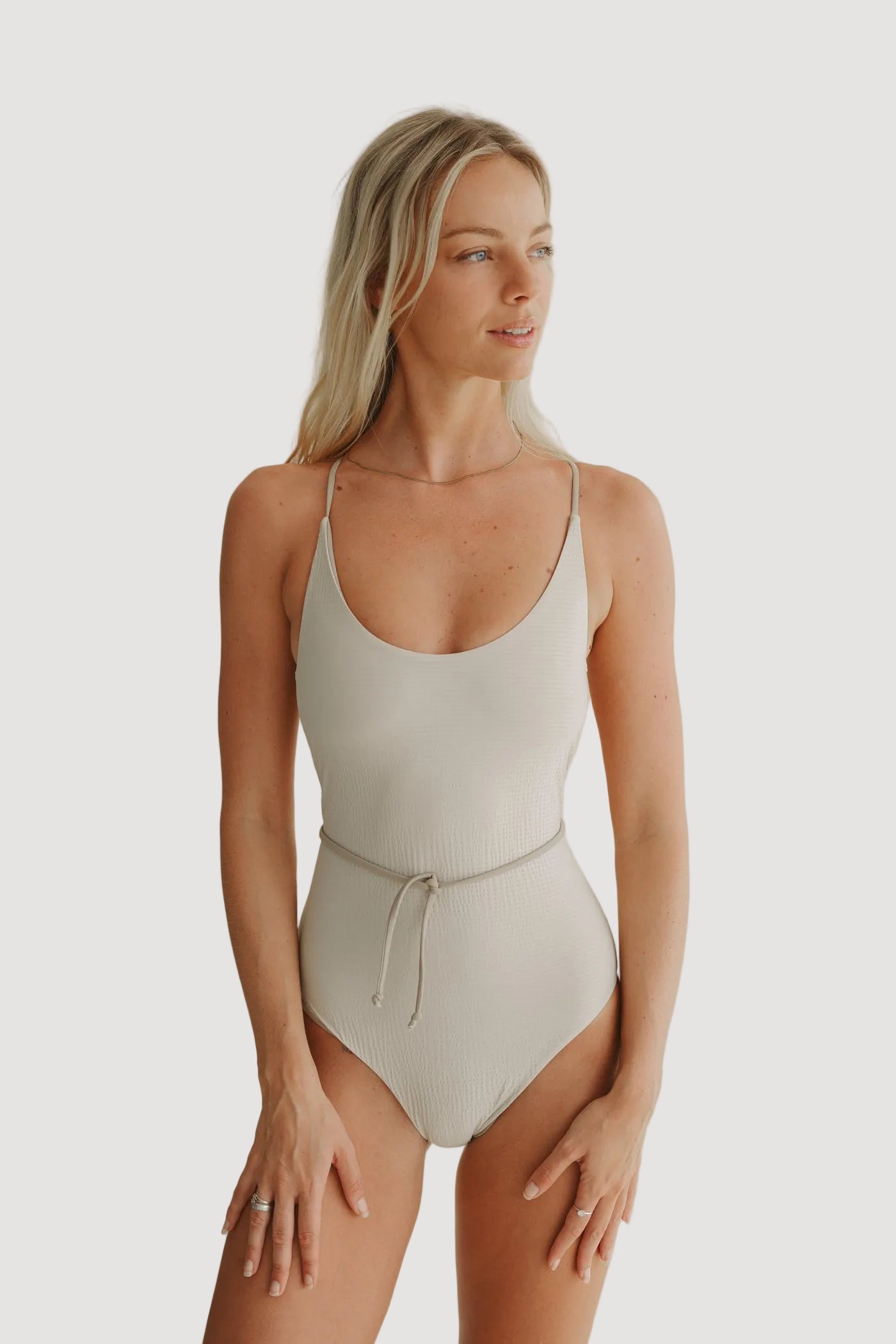 A female model wearing a beige one-piece swimsuit with adjustable straps and a V-neckline.