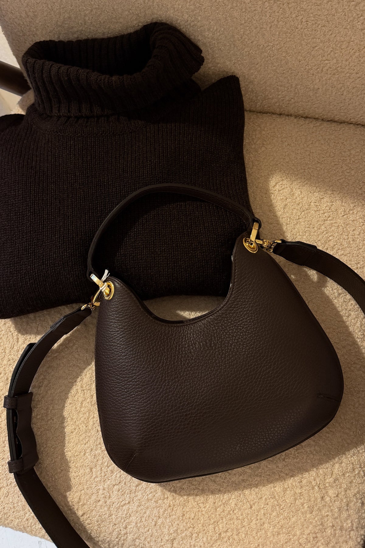 A dark brown pebbled leather crescent-shaped shoulder bag with gold-tone hardware, resting on a beige carpet next to a black knit sweater.
