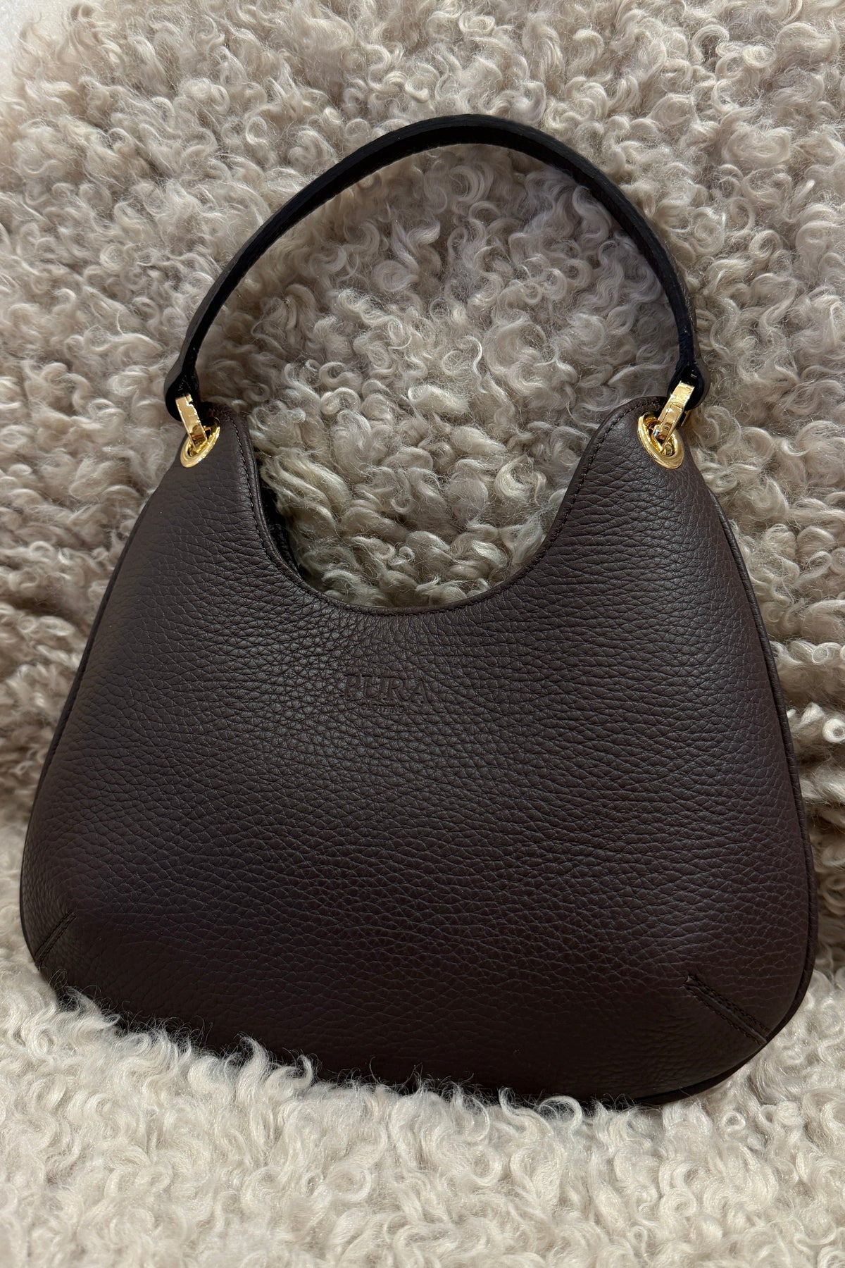 A dark brown pebbled leather shoulder bag with gold-tone hardware, resting on a white shaggy rug.