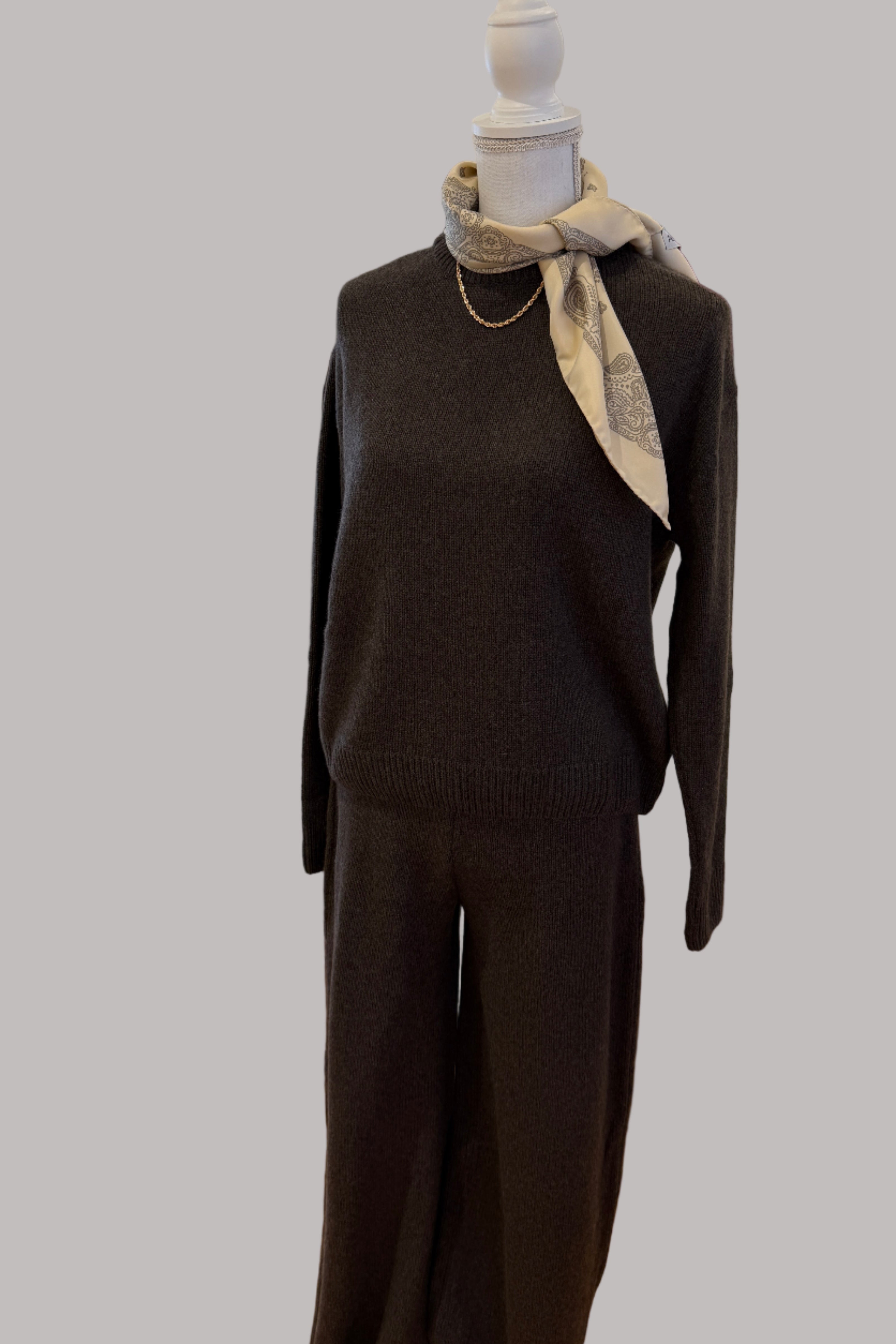 Mannequin wearing a dark outfit with a scarf in a room with boxes and another garment in the background.