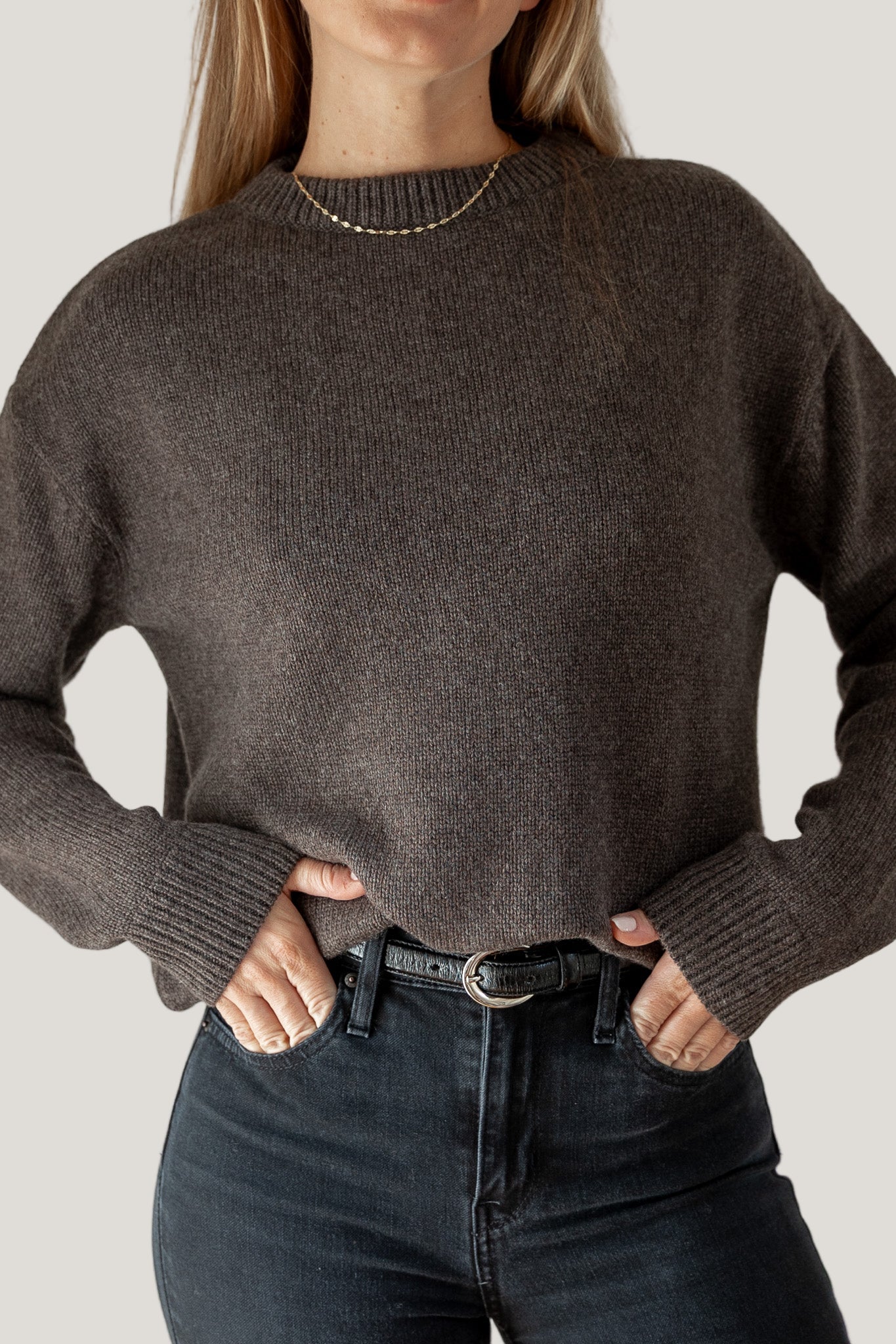 Person wearing a brown sweater and dark jeans against a neutral background