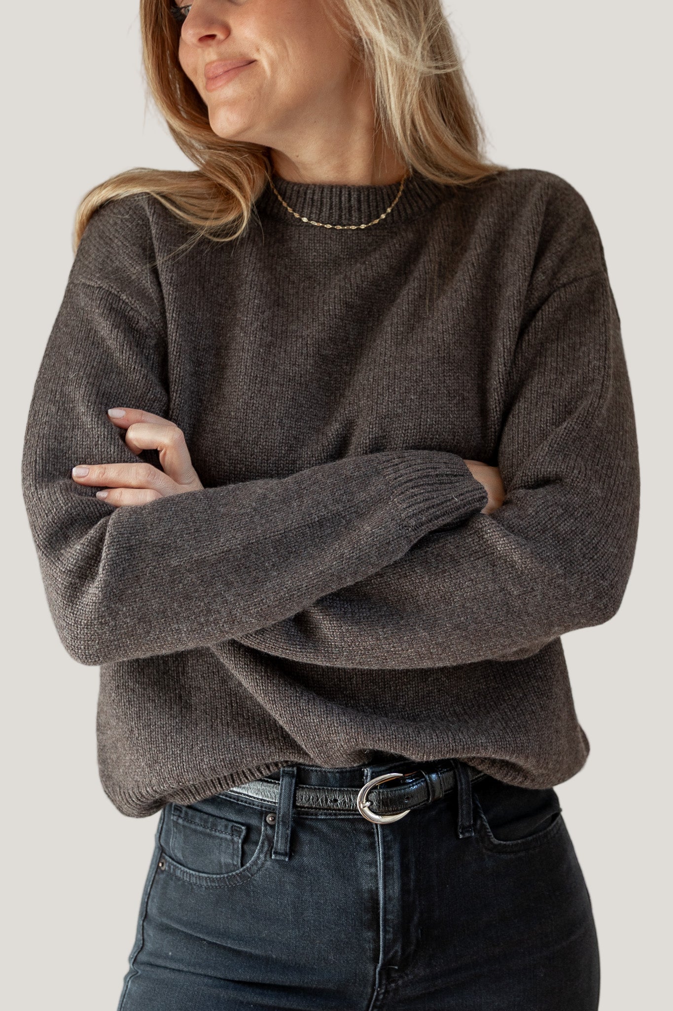Woman wearing a brown sweater and dark jeans against a plain background