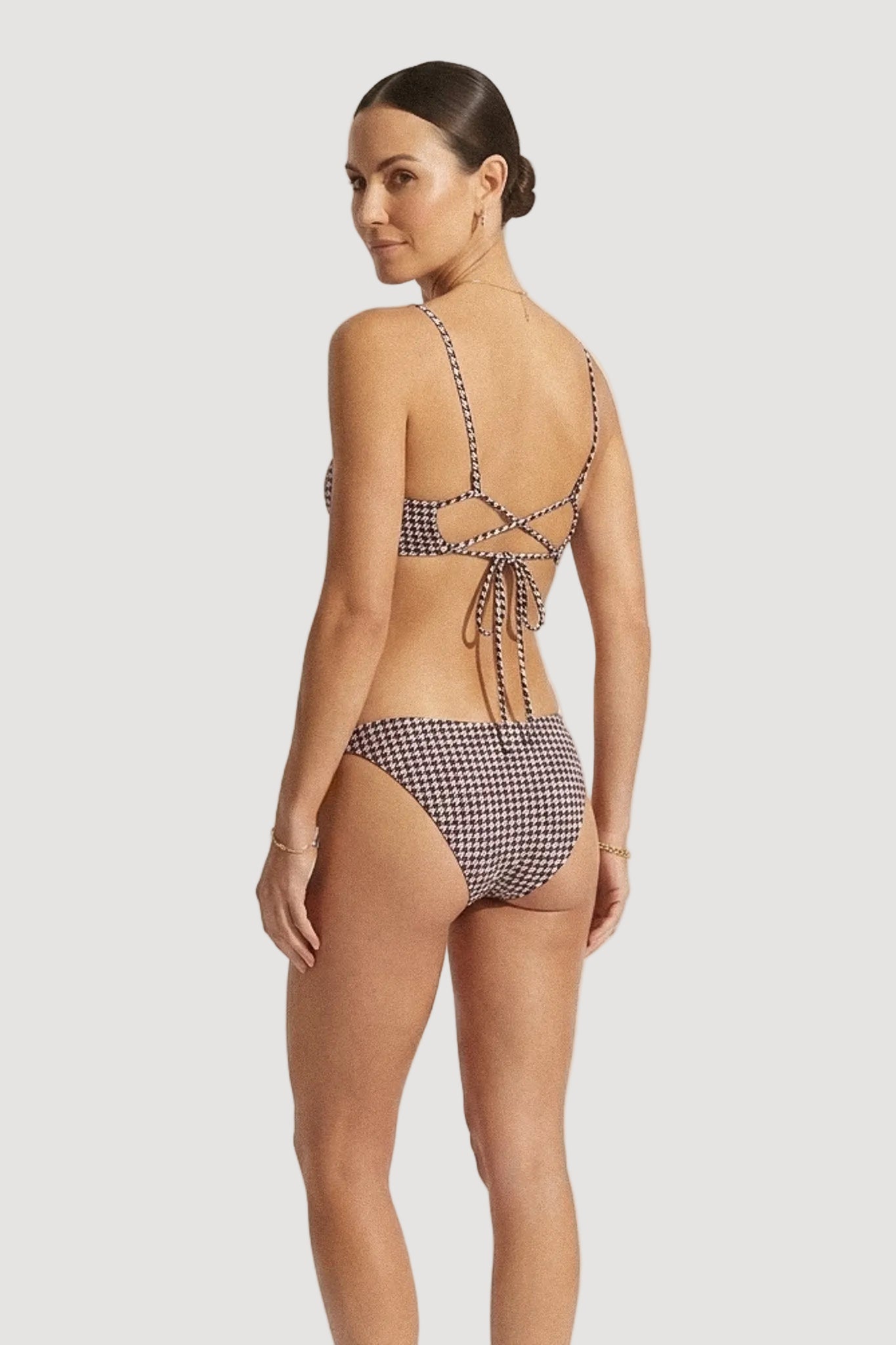 A woman modeling a two-piece bikini from the back, featuring a navy and beige gingham pattern with tie-back straps on a light gray studio background.