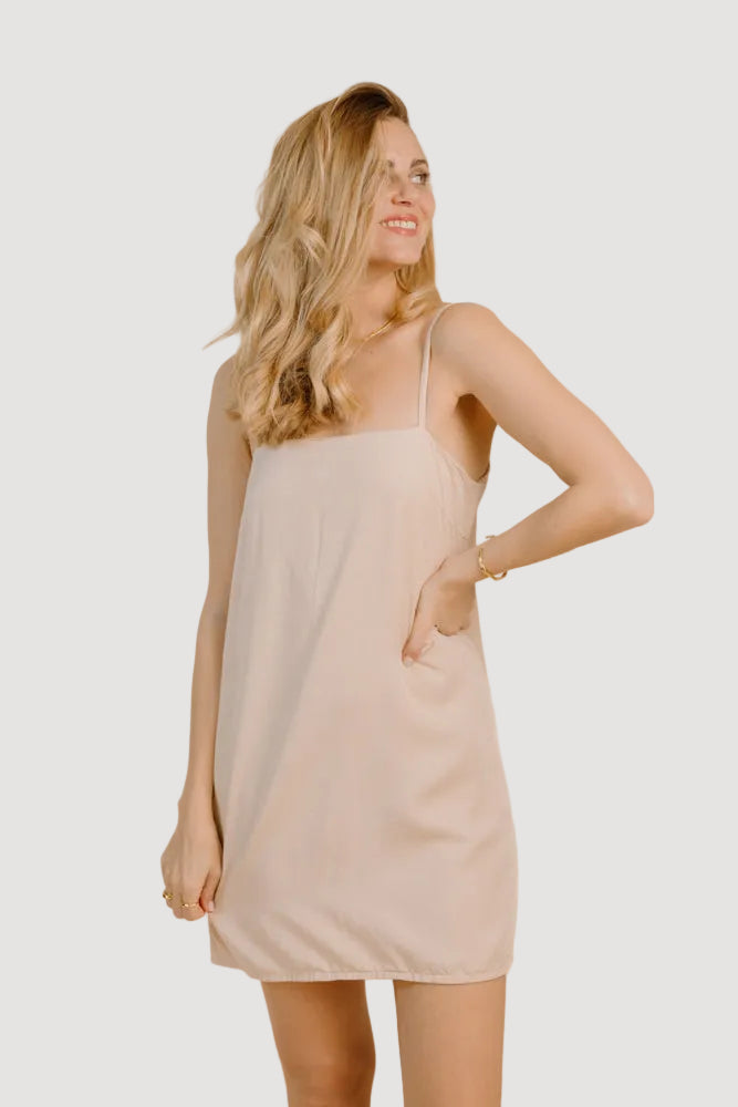 A female model wearing a beige, sleeveless, mini dress with adjustable spaghetti straps, standing against a plain background.
