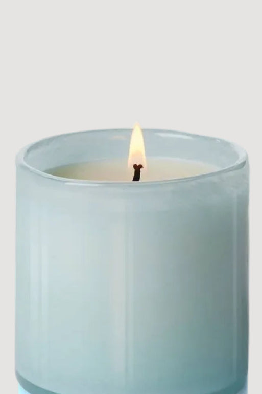 A marine-colored candle in a clear handblown art glass vessel with a lit wick, set against a white background.