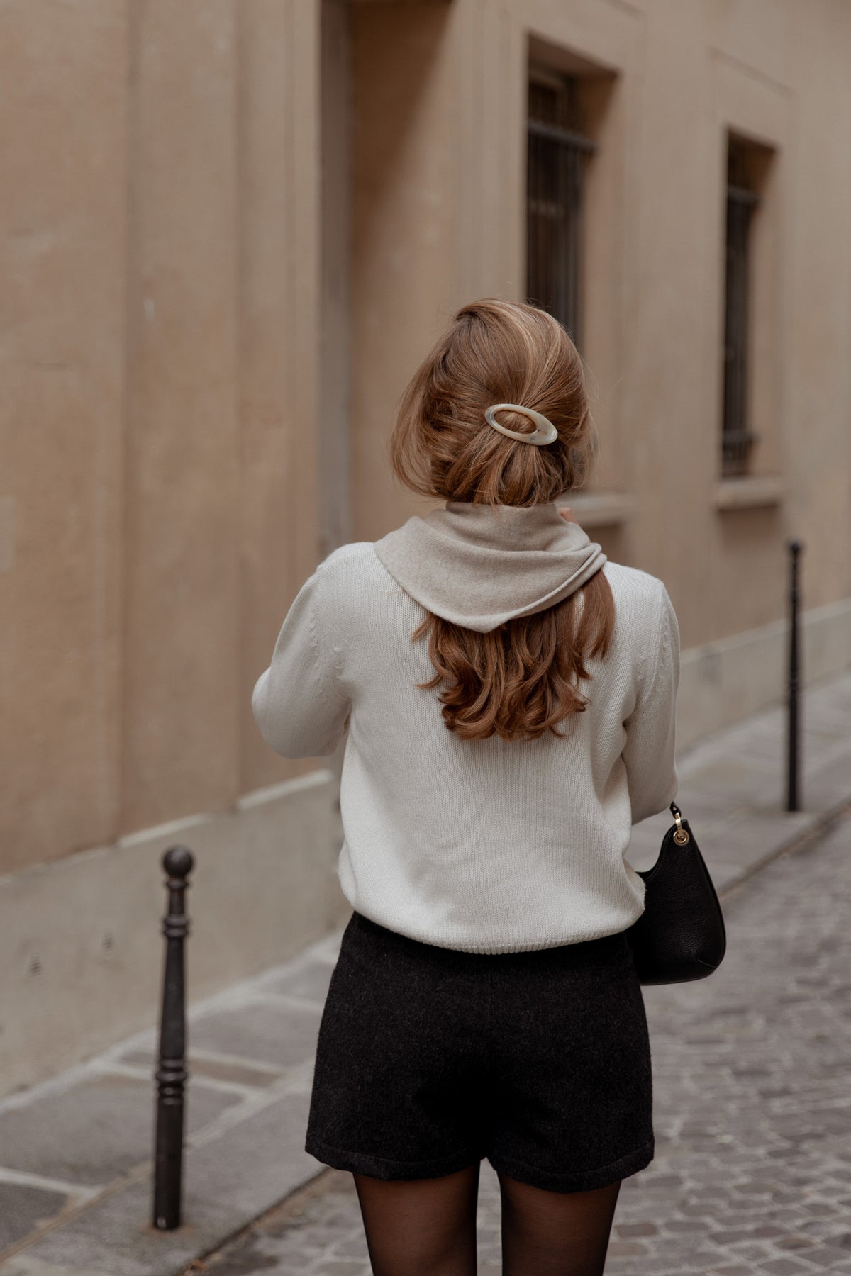 A woman viewed from behind wearing a cream knit sweater, black shorts, and black tights, carrying a black shoulder bag while walking on a cobblestone street.