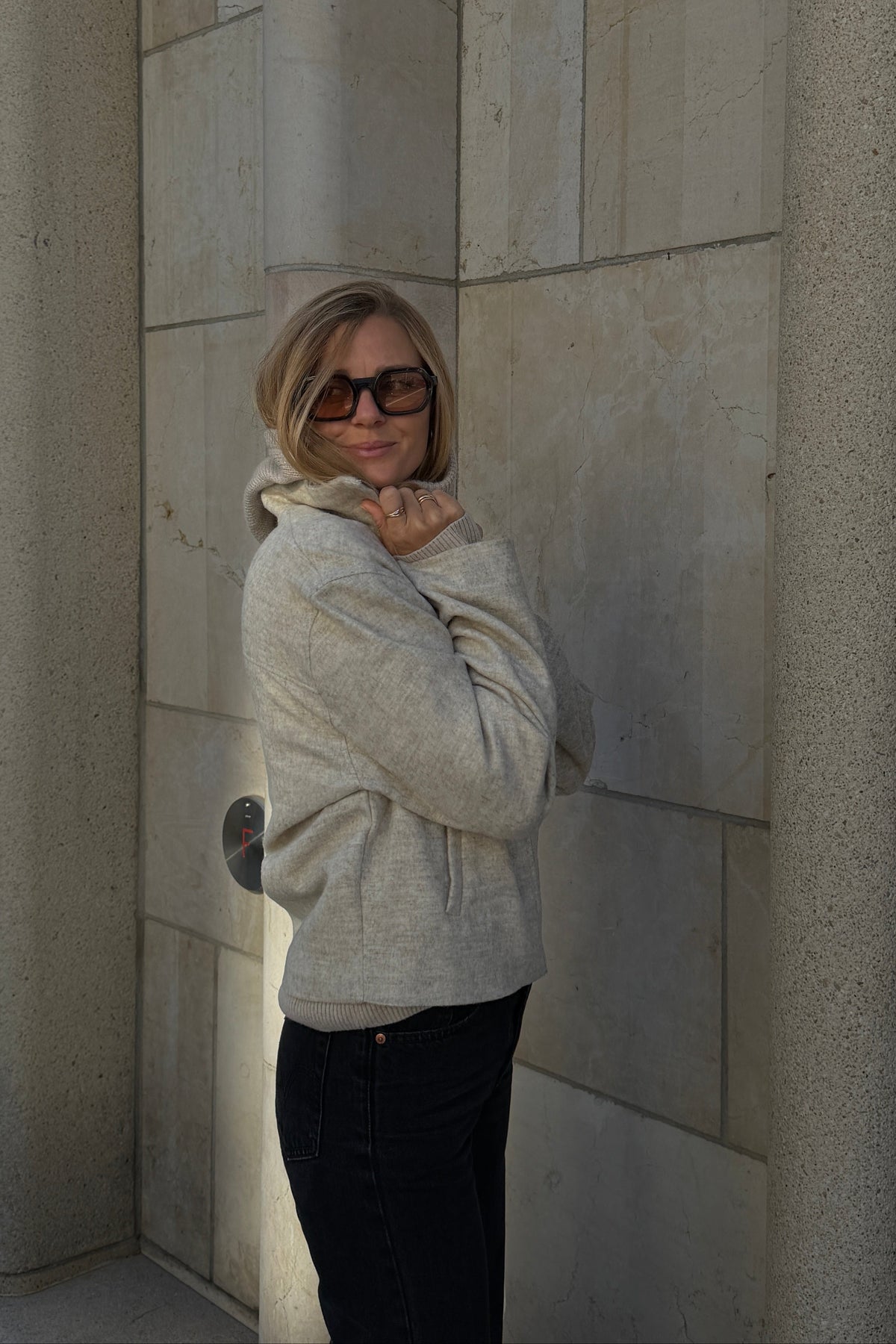 A woman with blonde hair and sunglasses stands in a stone-walled corridor wearing a light beige knit hoodie and dark pants.