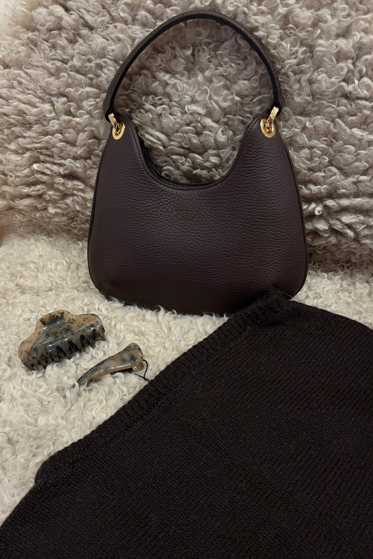 A dark brown pebbled leather shoulder bag with gold-tone hardware, displayed on a textured gray shag rug alongside a black knit sweater and two small accessories.