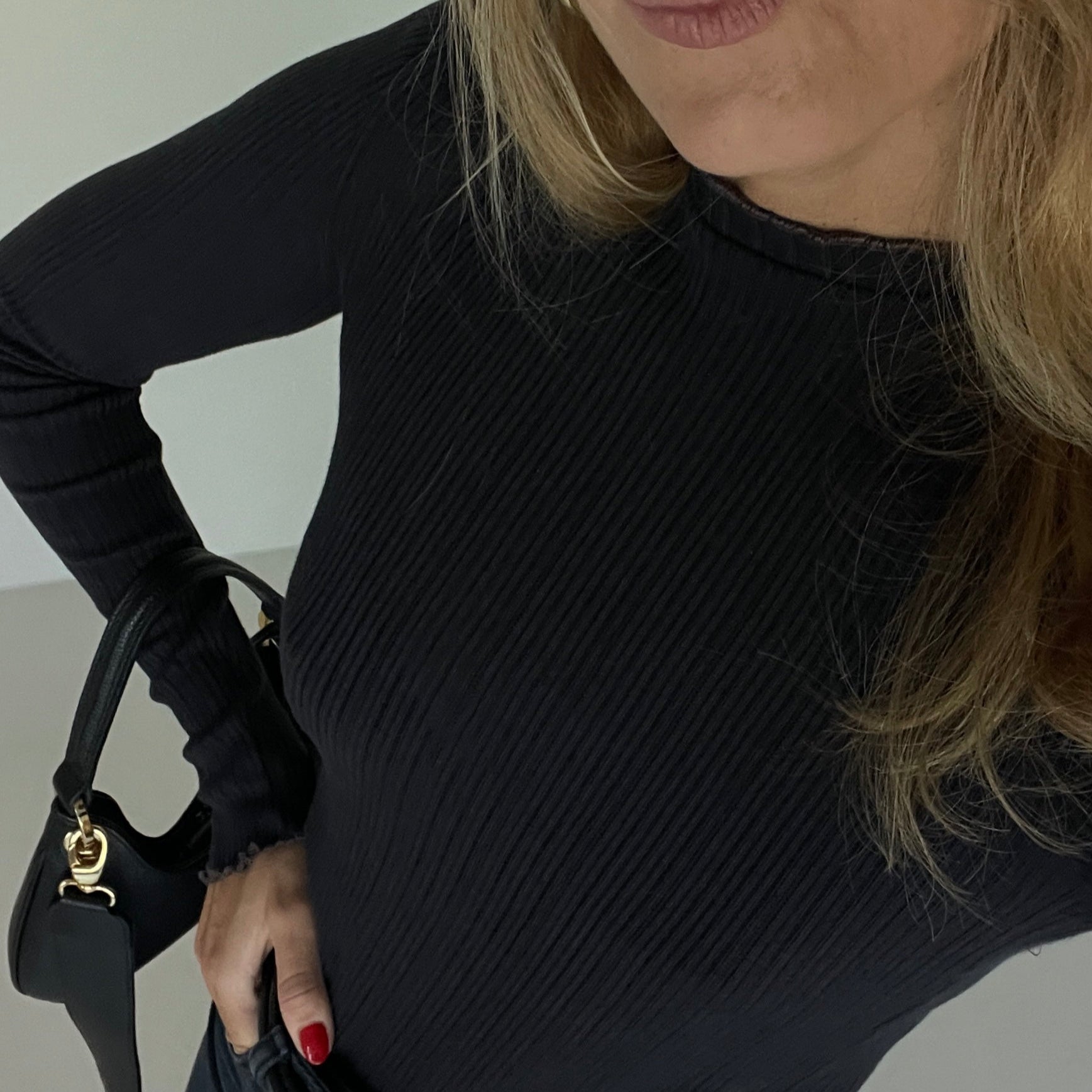 Woman wearing a black ribbed sweater and sunglasses, standing against a plain background.