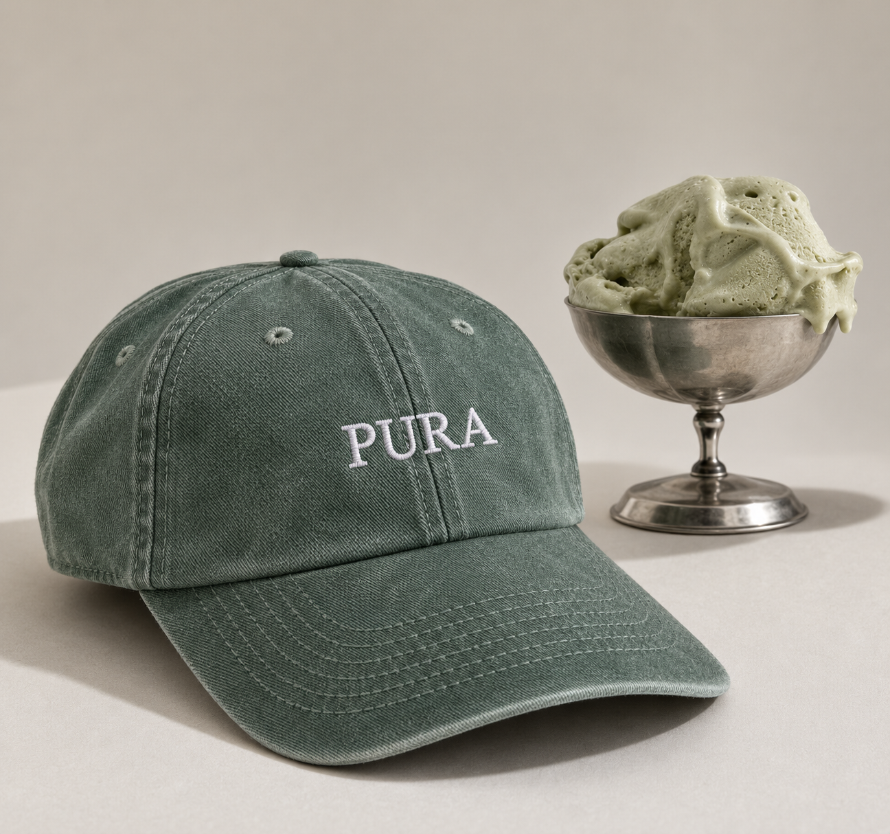 A dusty sage green cotton baseball cap with 'PURA' embroidered in white on the front, styled next to a silver pedestal bowl filled with mint green ice cream on a light beige background.