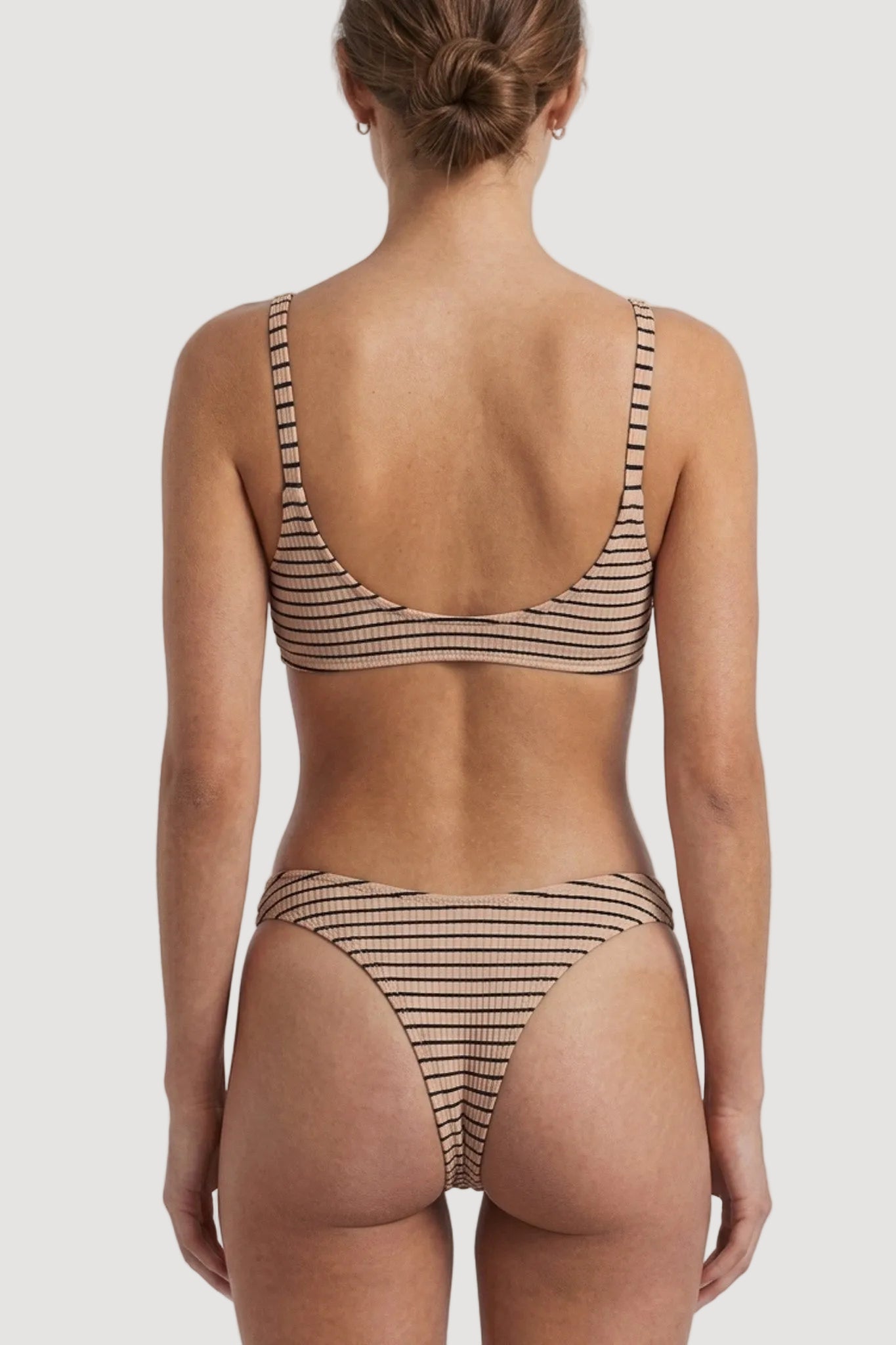 Rear view of a model wearing a two-piece bikini with thin horizontal brown stripes on a beige background, set against a solid light gray studio backdrop.