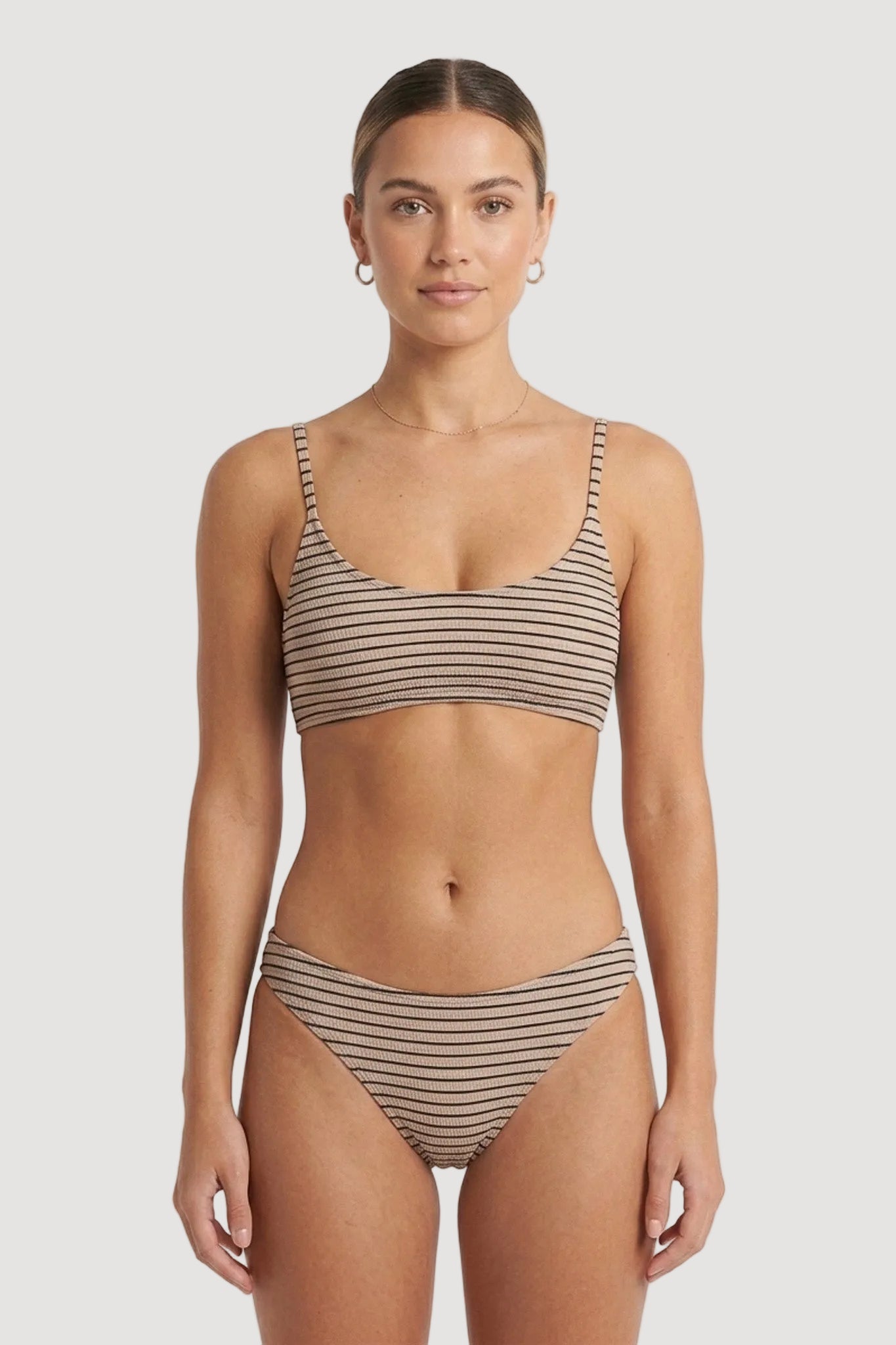 A woman modeling a two-piece bikini with thin horizontal stripes in beige and brown, photographed against a plain light gray studio background.
