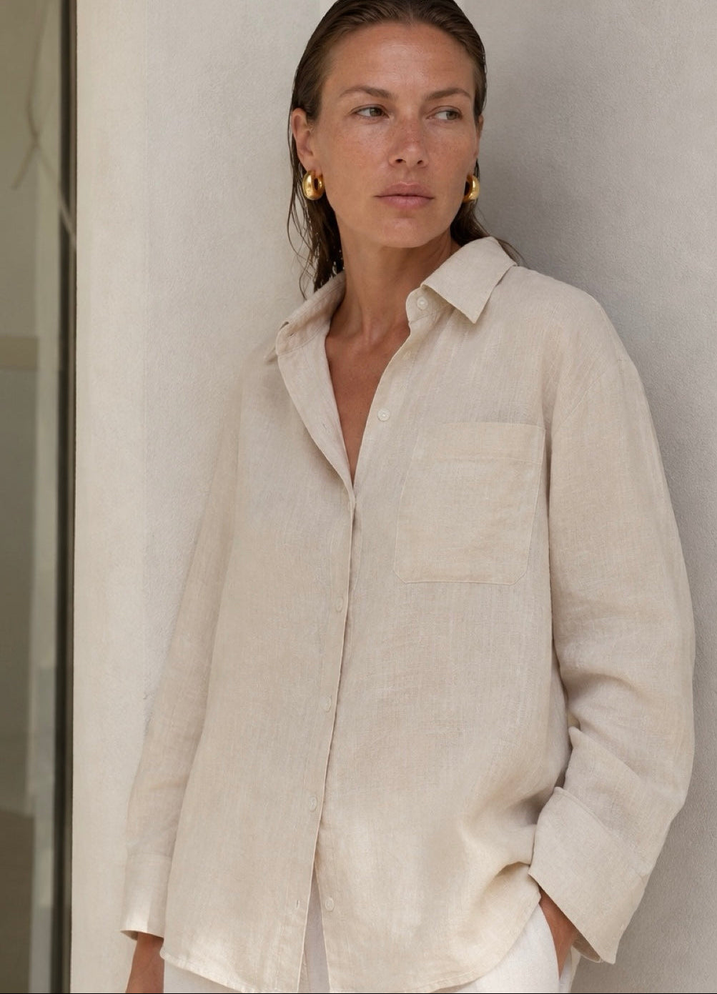 A woman wearing a light beige linen button-up shirt with a chest pocket, posed against a neutral textured wall. The brand logo 'PURA CLOTHING' is visible at the top of the image.